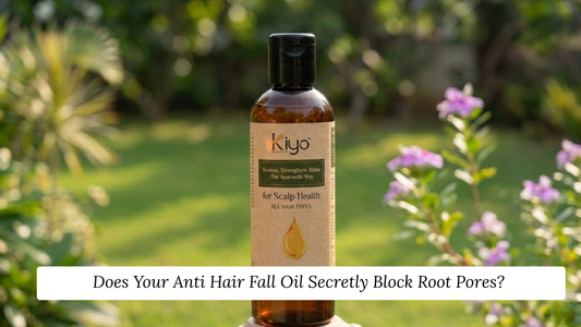 A 100ml amber glass bottle of Kiyo Beauty Ayurvedic Scalp Health Hair Oil centered against a soft, blurred green garden background with light pink flowers under warm natural daylight.